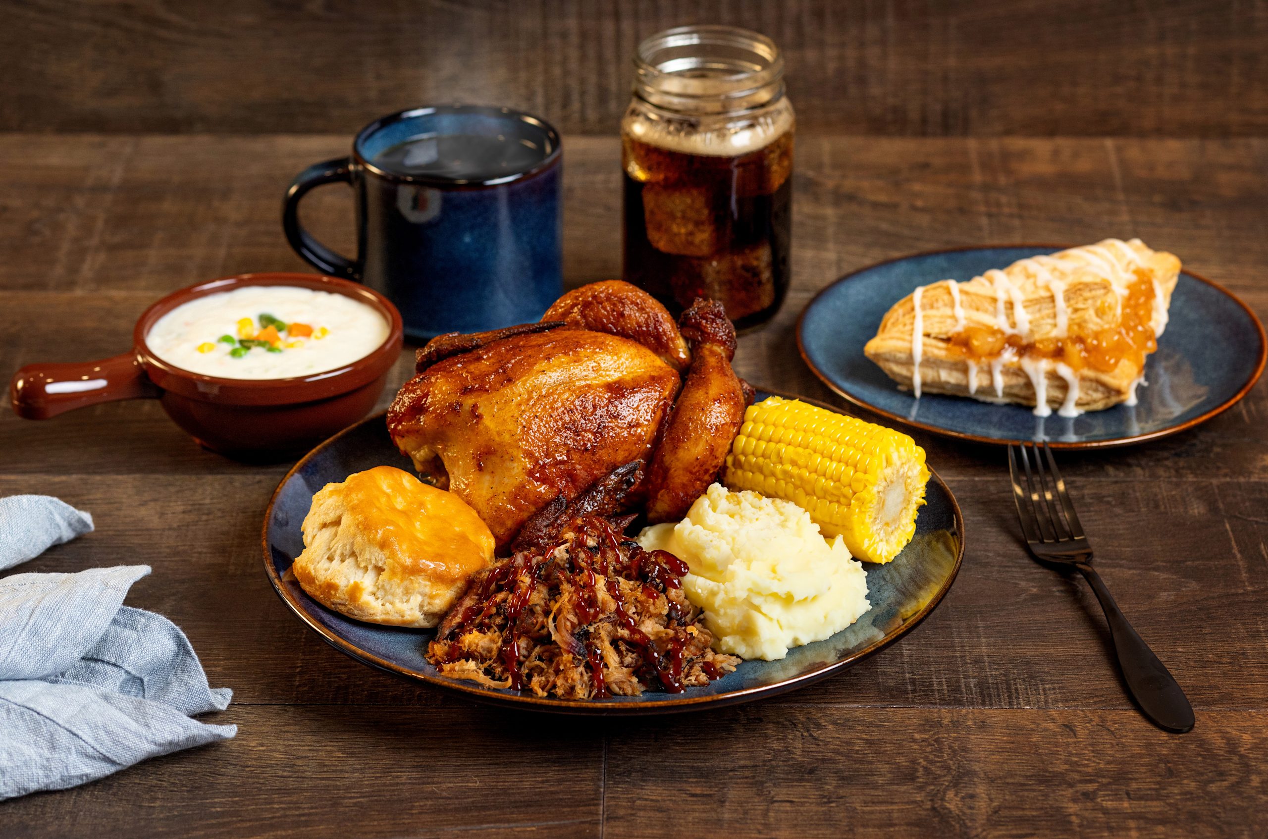 A roast chicken dinner with pulled pork, mashed potatoes, corn on the cob and a biscuit beside a bowl of soup and a fruit turnover on a dark wood table