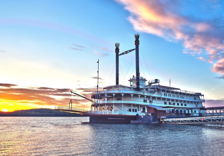 A riverboat with twin smokestacks moored at a lakeside dock at sunset