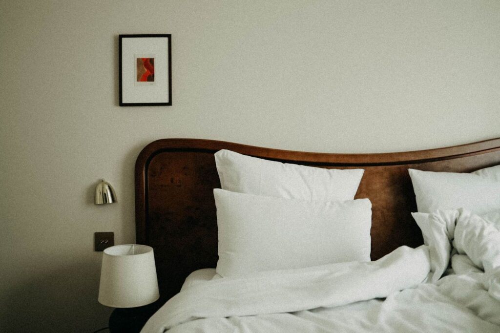 Hotel bed made up with white linen beneath a dark wood headboard and a shaded bedside lamp
