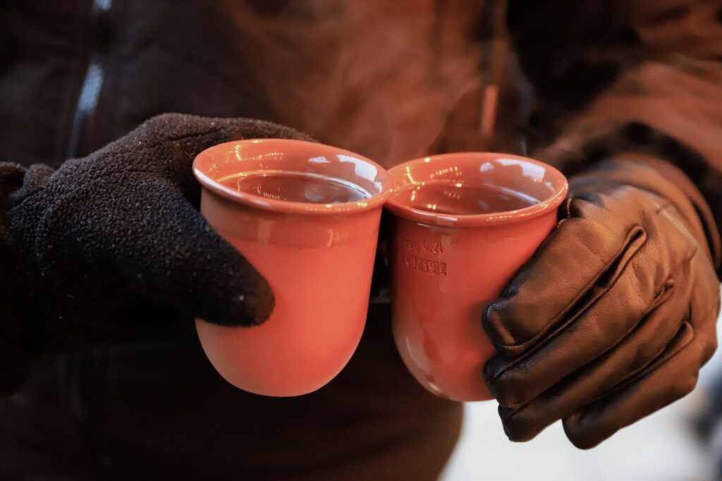 Two gloved hands clinking terracotta mugs of hot mulled wine at a Christmas market