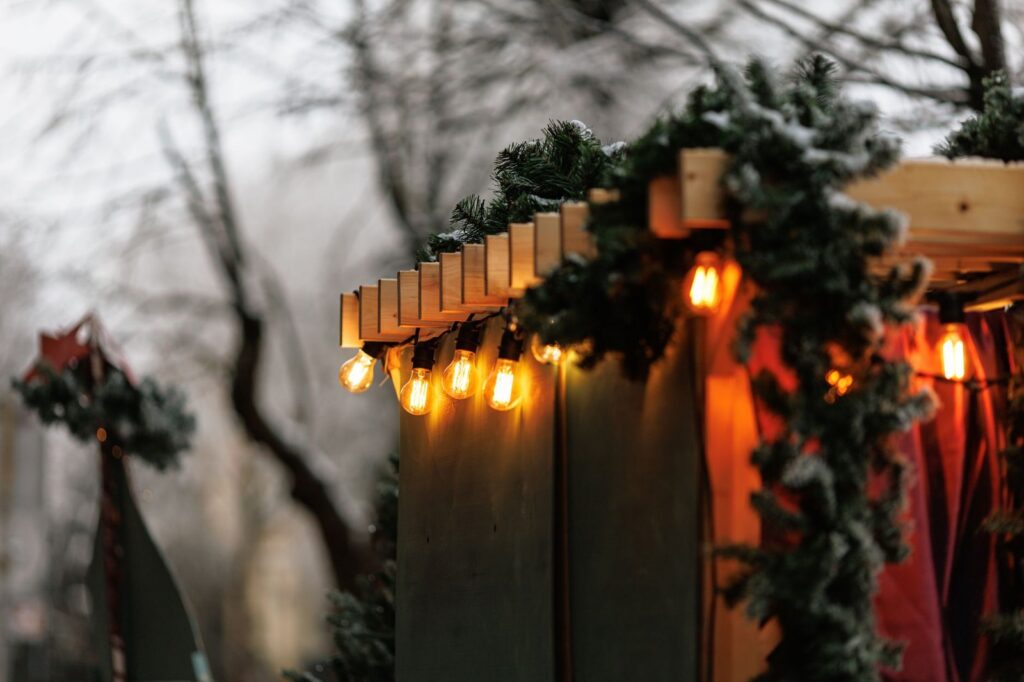 Warm bulb lights and a snowy evergreen garland strung along the wooden roofline of a Christmas market stall