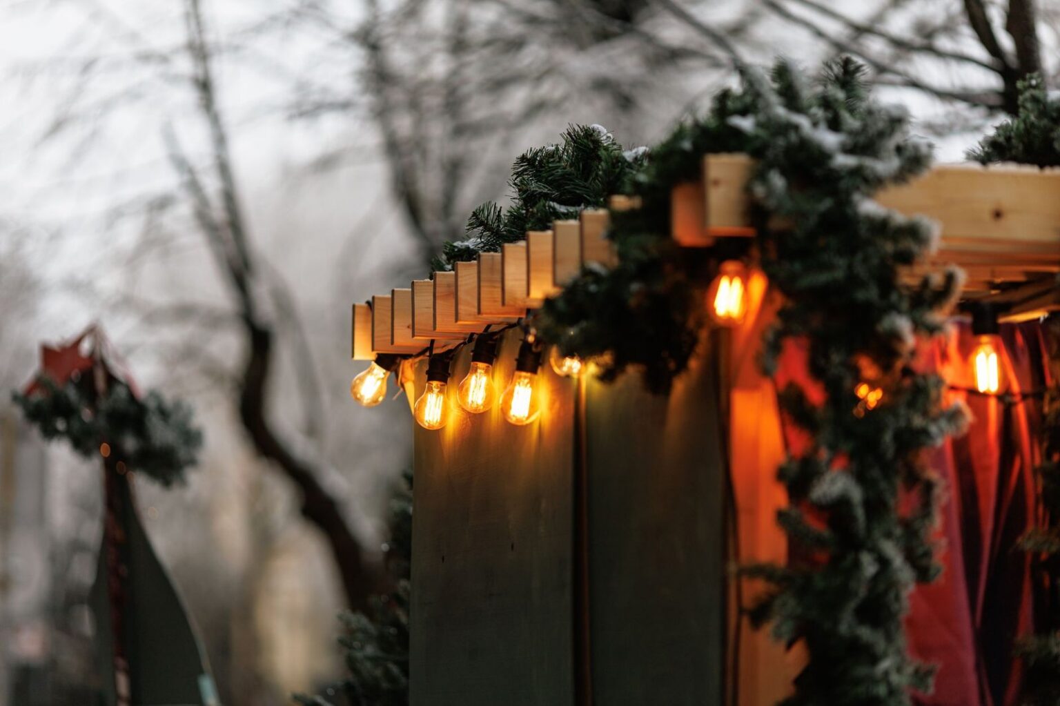 Warm bulb lights and a snowy evergreen garland strung along the wooden roofline of a Christmas market stall