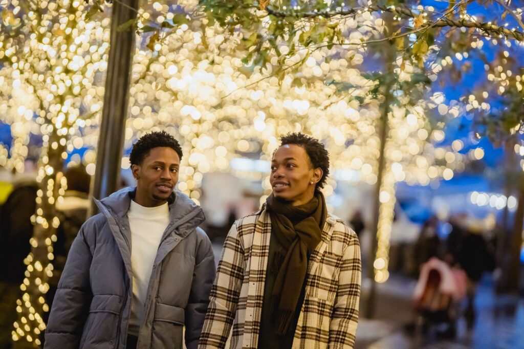 Two people in winter coats walking under trees strung with illuminated garlands in the evening