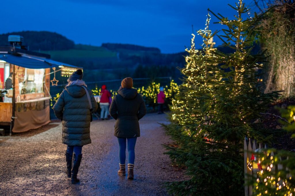Two visitors in winter coats walking a gravel path past a lit food hut and illuminated trees at an outdoor Christmas market