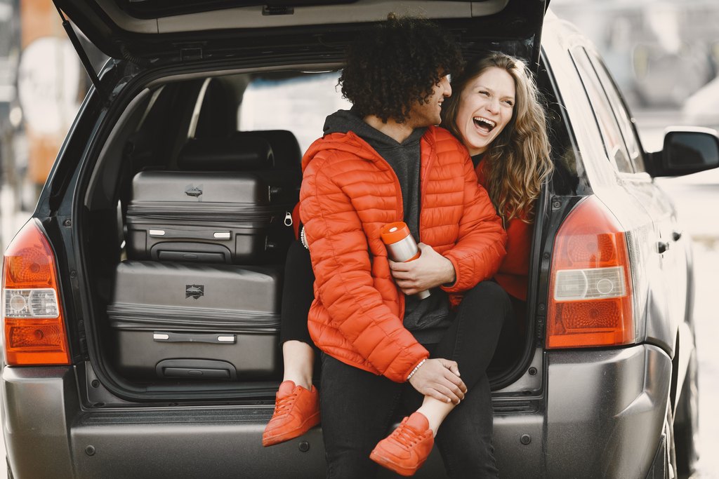 Two travelers laughing while sitting in the open trunk of a car packed with suitcases