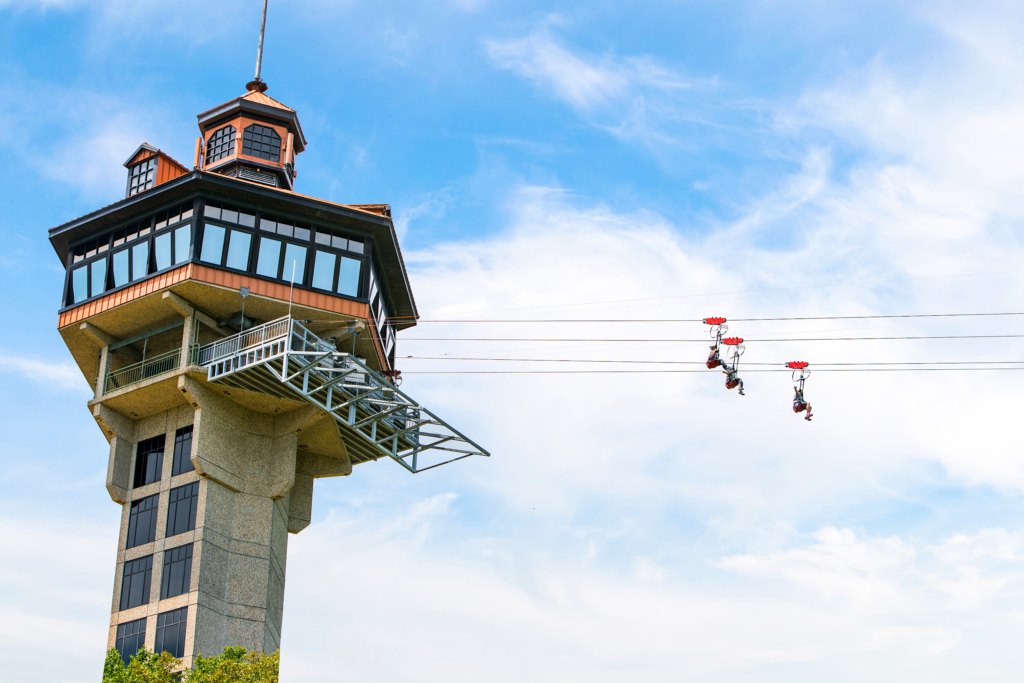 Two riders descending a long zipline cable from a tall observation tower against a blue sky