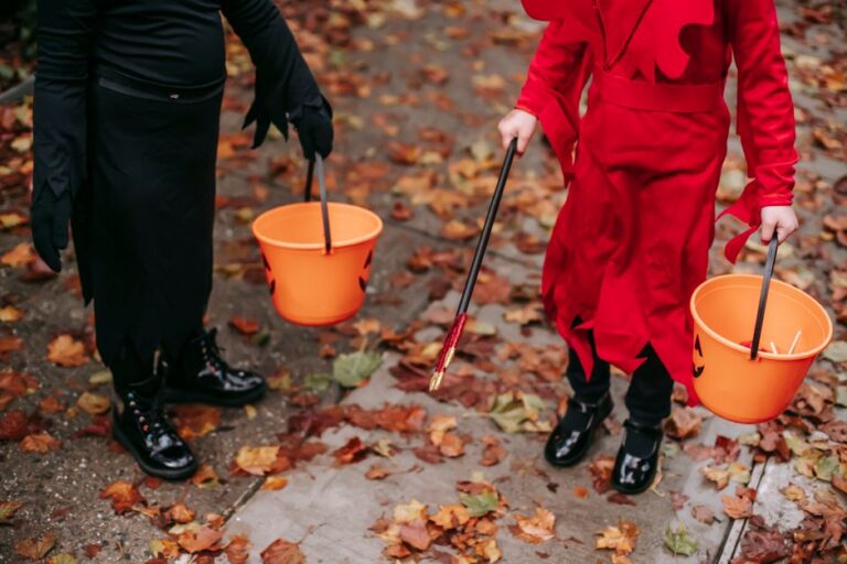 Two children in black and red Halloween costumes carrying orange jack-o-lantern buckets on a leaf-covered path, shown from the waist down